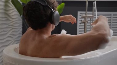 Beyaz tenli seksi, çıplak, dinlenen adam köpüklü küvetten zevk alıyor sıcak su köpüğü olan erkek, banyoda dinleniyor kulaklıkla müzik dinliyor, kulaklıkla dans ediyor.