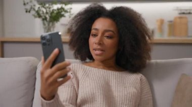Afro-Amerikalı kadın, kaygısız, etnik bayan ev sahibi kanepe üzerinde gülümsüyor video konuşması, akıllı telefon, ev telefonuyla uzaktan iletişim kuruyor.