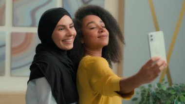 Çeşitli çok ırklı iş kadınları mutlu mutlu kız iş ortakları Arap müslüman kadın tesettüre ve afro-amerikan kadınlara gülümseyen selfie pozu veriyorlar ofisteki cep telefonu kamerasında.