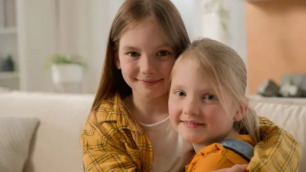 两个可爱的 面带微笑的白俄罗斯孩子们 小女孩 姐姐们 朋友们拥抱拥抱在一起 抱着拥抱在家里沙发上 在客厅里摆出温馨的姿势 和家人团聚在一起 — 图库照片