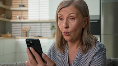 60 'lı yaşlarda, mutlu, beyaz bir kadın ev bankasındaki koltukta cep telefonu kullanarak iyi haberler okuyor. Kadın emeklinin akıllı telefondan online kazanmasını hayrete düşürüyor. Piyango zaferini kutluyor.