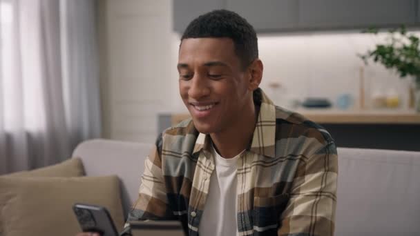 アフリカ系アメリカ人男性バイヤーケアフリー男性エスニック男性クライアント顧客笑顔で興奮した注文オンライン配信アプリのバンキング決済ショッピング クレジットバンクカードと自宅で携帯電話 — ストック動画