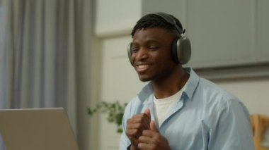 Mutlu, gülümseyen, Afro-Amerikan erkek adam evde müzik dinlemekten zevk alıyor. Kablosuz kulaklıkla kulaklık takıyor, internette dizüstü bilgisayarla dans ediyor, dans ediyor, el dansı yapıyor, özgürlük dansı yapıyor.