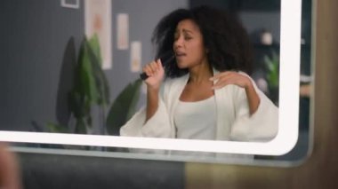 Banyoda eğlenen mutlu kız dans eden Afro-Amerikalı kadın kıvırcık saçlı kadın evde ayna güzellik dansına bakıyor mikrofon gibi saç fırçasında şarkı söylüyormuş gibi yapıyor.