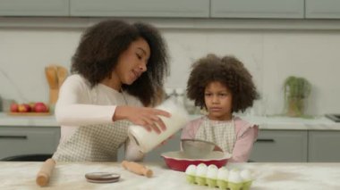 Afro-Amerikalı anne, küçük kızına mutfakta birlikte yemek pişirmesinde yardım ediyor. Mutlu bir aile, pasta malzemesi, pasta pişiren bir kız çocuğu, elek, anne, un döküyor. Yüksek