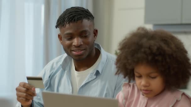 ハッピーアフリカ系アメリカ人の家族ショッピング 自宅のキッチンでオンラインコンピュータを購入 母親父親は 小さな子供の少女娘を教えるクレジットバンクカードを保持しています 製品を購入する 食品配達ラップトップ購入 — ストック動画