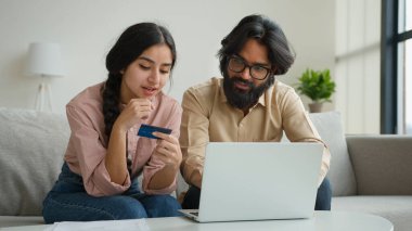 Arap Hintli Müslüman çift, oturma odasındaki koltukta oturuyor. Kredi kartı şifresi, bilgisayar bankacılığı uygulamasında geçiyor. Çevrimiçi çok ırklı kadın ve internet alışverişi yapıyor.