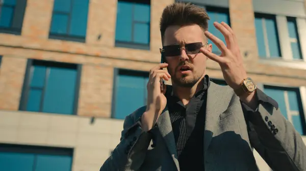 Annoyed businessman in sunglasses talk on phone angry displeased man entrepreneur leader solve business problems using smartphone near outdoors office furious nervous stressed customer arguing crisis