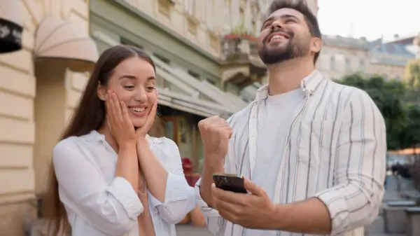 Caucasian happy couple enjoy excited man woman shocked lucky success victory mobile phone smartphone reading message good news amazed surprised open mouth city outdoors urban gen z win wonder awesome