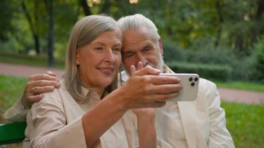 Mutlu Kafkasyalı büyükanne ve büyükbabası emekli çift erkek kadın dışarıda cep telefonu teknolojisi videosu öğrenirken eğleniyor. Yaşlı çift el sallıyor. Akıllı telefon kullanıyor. Açık hava bankında oturuyor.