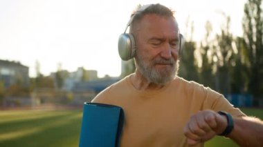 Neşeli yaşlı adam elinde spor minderiyle stadyuma gidiyor. Müzik kulaklıkları dinliyor. Kol saati eğitimine bakıyor.
