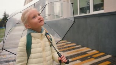 Küçük liseli kız sokakta yürüyor. Binalara bakıyor. Çocuk yuvası, hafta sonu dersleri, ilkokul gezintileri, şemsiye yağmuru çocuğu.