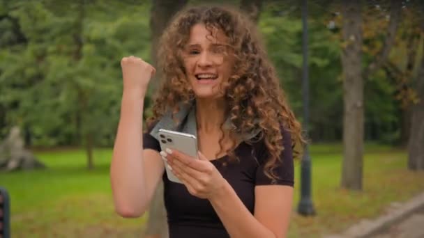 ハッピー驚いた白人女性若いガールブロガーインフルエンサーモバイルスクリーンウィン賞を見て トレーニング目標を達成 スマートフォンアプリでスポーツトレーニングの良い結果 携帯電話の喜び都市公園 — ストック動画