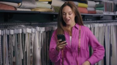 Kafkasyalı kadın mutlu, heyecanlı kadın moda tasarımcısı, terzi, cep telefonu şoku kullanıyor. Şaşırtıcı bir sürpriz, akıllı telefon tebessümü, kumaş dükkanında kahkahalar.