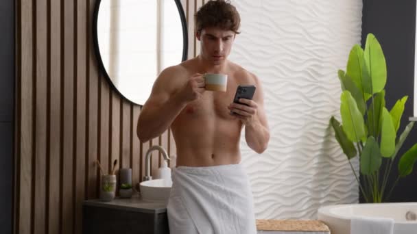 白人リラックスした裸の男は朝のルーチン読書ニュースで紅茶を飲む スマートフォンのソーシャルメディアで 浴室でホットコーヒーを飲む携帯電話でブラウジング 男は驚いた衝撃ショッキングなスピッキング飲料 — ストック動画