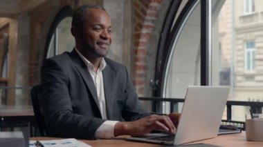 Afro-Amerikalı orta yaşlı girişimci gülerek mutlu gülümsüyor dizüstü bilgisayarlı, etnik iş adamı ofis işadamı gülümsüyor bilgisayarla internet üzerinden iş görüşmesi yapan erkek müşteri ile sohbet ediyor