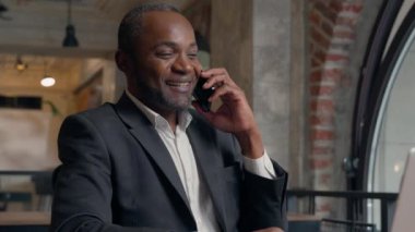 Mutlu gülüşler Afro-Amerikan pozitif etnik erkek iş adamı işveren gülümsemesi hoş sohbetler akıllı telefon konuşması iş adamı ofiste dizüstü bilgisayar yazan erkek.