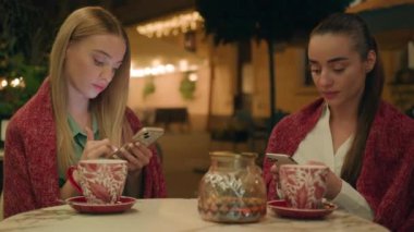 İki kadın, kafeterya restoranının dışında oturan en iyi arkadaşlar cep telefonu mesajlaşmaları, sohbet mesajlaşmaları, internet üzerinden sıcak çay mesafesinde mesajlaşmalar, bağımlı sanal hayat.