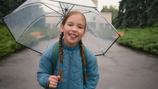 快乐的小女孩看着相机露出舌头鬼鬼祟祟的表情女学生女儿溺爱着孩子们孩子们快乐的雨天周末精力充沛的快乐的公园城市 — 图库视频影像