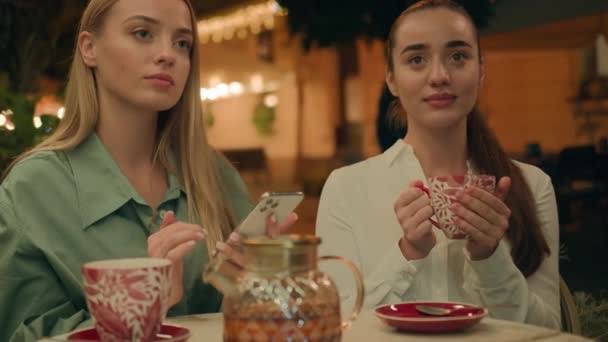 若いヨーロッパの白人女性カフェレストランで2人のガールフレンドが一緒に紅茶コーヒーを飲む表情グリムースは カップスマートフォン電話を保持する嫌悪を無視する外の刺激反応都市を獲得します — ストック動画