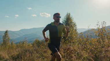 Atletik Kafkasyalı erkek sporcu erkek spor giyim gözlüklü koşucu vadi yürüyüşü yapıyor dağlara tırmanıyor açık havada yeşil tepe tırmanıyor macera macerası yapıyor sağlıklı yaşam tarzı egzersizleri yapıyor.