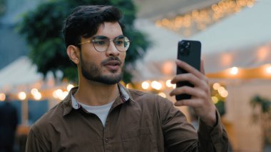 Yakışıklı, Hintli, Arap, etnik, erkek, öğrenci, erkek, vlogger, iş adamı, vlogger, video kaydı yapan kişi. Cihaz cep telefonu uygulaması kullanarak, akşam kenti sokaklarında yürüyor.