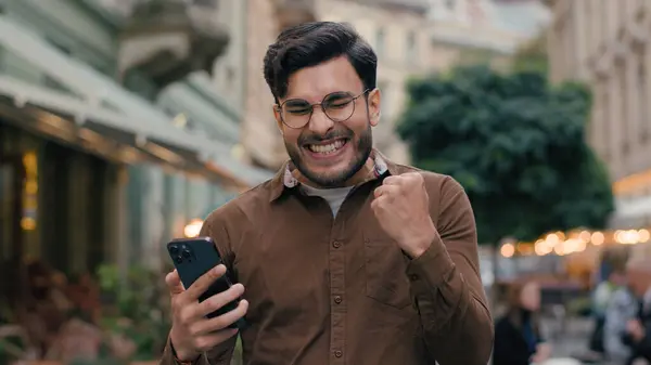 快乐而有希望的印度裔男人生意人男人拿着手机年轻的学生希望祈祷中指的人是城市街道外的尖叫胜利姿态 图库照片