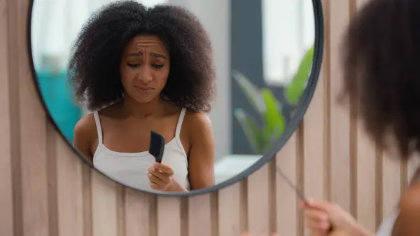 灰心丧气的非洲裔美国女人遇到了麻烦 卷发看着镜子看浴室的美容美发例行公事 准备用梳子梳理卷曲纠结的卷发 图库照片