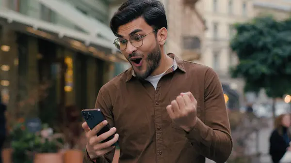 快乐而有希望的印度裔男人生意人男人拿着手机年轻的学生希望祈祷中指的人是城市街道外的尖叫胜利姿态 免版税图库照片
