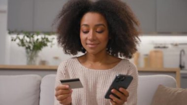 Mutlu gülümseyen Afrikalı Amerikalı kadın cep telefonu ve kredi kartı alışverişini internet üzerinden yapıyor. Akıllı telefondan ödeme alıyor. Etnik kadın alışverişçi ev mutfağında dijital bankacılık kullanıyor.
