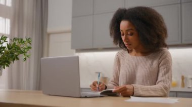 Afro-Amerikalı etnik kız öğrenci evde dizüstü bilgisayarla çalışıyor. Uzak karantina eğitiminde çalışıyor. Serbest çalışan kadın işkadını mutfak masası bilgisayarına notlar yazıyor.