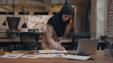 Müslüman İslami kadın tesettüre girmiş dizüstü bilgisayar projesi analizi planlayan, gülümseyen kadın iş kadını masada cep telefonu kulübesinde konuşma notları yazan, ofisteki akıllı telefon konuşmalarını yapan.