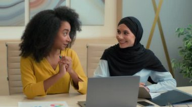 Çok ırklı iş arkadaşları kadın iş kadınları Afrikalı Amerikalı işveren Arap müslüman islami yönetici tesettüre bağlı mutlu ofis çalışanları birlikte gülerek dizüstü bilgisayardaki projeyi tartıştılar