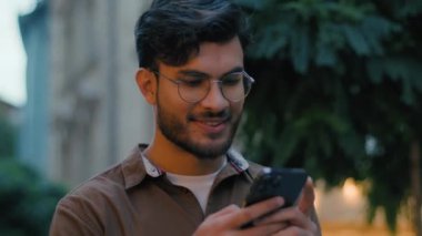 Yakın plan başarılı Hintli Arap etnik erkek erkek öğrenci genci iş adamı mutlu akıllı telefon kullanıcısı kayan cep telefonu aygıtı ile sohbet ediyor online uygulama uygulaması sohbet gece şehir caddesi açık