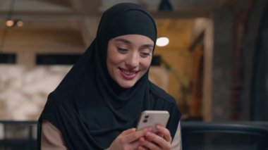 Müslüman kız Arap İslami tesettürlü mutlu kadın iş kadını ofis yöneticisi cep telefonu daktilo mesajları kullanarak akıllı telefon sohbeti mesajları yazarak gülümsüyor.