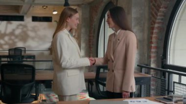 Two happy smiling friendly Caucasian businesswomen girls female business women partners friends coworkers colleagues managers executives in office meeting handshaking greeting talking conversation