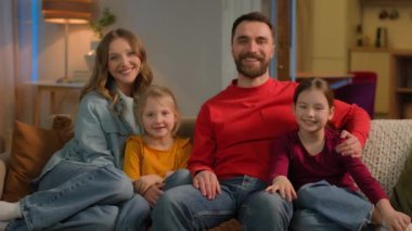 Caucasian family bonding portrait happy parents kids on couch sofa weekend cheerful mom dad little girls daughters siblings sisters children greeting waving hands hello smile affectionate home owners