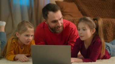 Caucasian happy family father kids daughters little girls lying on floor home night order delivery buying online shopping laptop computer playing game watch cartoons laugh dad help e-learning children
