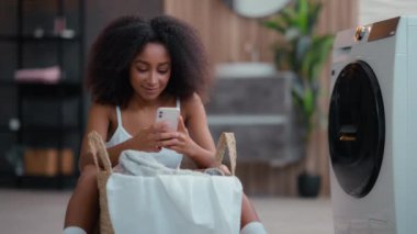 Afro-Amerikan kadın ev hanımı bayan mutlu mutlu gülümseyen kaygısız sohbet sosyal medya mesajı cep telefonu gülümsemesi kullanarak akıllı telefondan mesaj atarak çamaşır sepeti çamaşır makinesiyle yerde oturuyor.