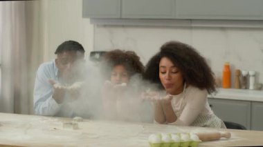 Afro-amerikan mutlu gülümseyen mutlu aile babası baba anne anne küçük kız evlat edinilmiş çocuk kız birlikte mutfakta un üfleme tozu pişiriyorlar kahkaha atarak neşe oyunu oynuyorlar