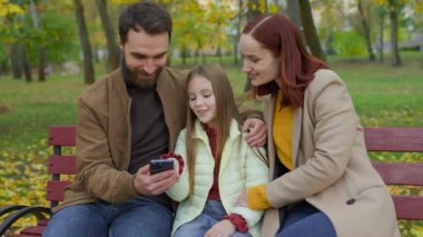 Beyaz, mutlu, gülümseyen bir aile haftasonu tatilini şehir banklarında, sonbahar parkında cep telefonlarını kullanarak birlikte gülümseyerek internet üzerinden alışveriş yaparak, akıllı telefon alışverişi yaparak, küçük kız, kız, çocuk, anne, baba...