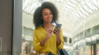 Mutlu Afro-Amerikalı kadın alışveriş merkezinde dikiliyor, alışveriş mağazalarında moda indirimi yapıyor. Kara Cuma 'da yeni şeyler alıyor. Gülümsüyor, mesajlaşıyor, sohbet ediyor, akıllı telefonlara bağlanıyor, web sitelerine bağlanıyor.