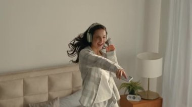 Kulaklıklı mutlu bir kadın yatak odasından zıplıyor en sevdiği şarkı müzik dinliyor, cep telefonu dinliyor, akıllı telefon kızı, teknik hareket yapıyor, dijital aletin keyfini çıkarıyor ve dansçıyı kutluyor.