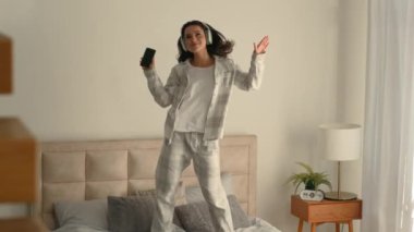 Yatak odasında zıplayan kadın ev kulaklıkları müzik dinliyor hafta sonu şarkıları söylüyor mutlu kadın cep telefonu teknolojisi çalma listesi enerji hareketli hareketli trambolinin tadını çıkarın