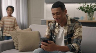 Neşeli Afro-Amerikalı etnik adam online cep telefonu oyunu oynamaktan zevk alıyor. Küçük çocuk çalışıyor mutlu çocuk babasının dikkatini dağıtıyor. Akıllı telefon bağımlılığı.