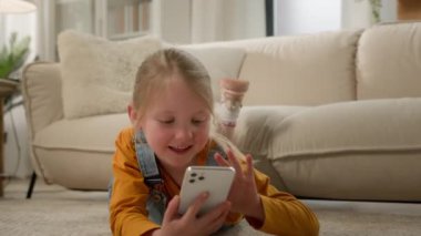 Mutlu gülen, kaygısız, küçük beyaz kız liseli kız, yerde yatıp oyun oynuyor cep telefonlu akıllı telefon kızı, sosyal medya evinde yazarak sohbet ediyor. Aygıtlar aşırı kullanılıyor