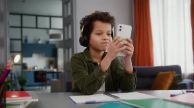 Kulaklıklı komik Afrikalı Amerikalı çoklu-ırkçı çocuk. Çocuk öğrenci evde eğitim görüyor. Dinlenmek için cep telefonu ses uygulaması kullanıyor.