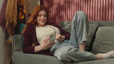 Kafkasyalı kızıl saçlı kadın ev sahibi bağımlı cep telefonu kullanıcısı mutlu kızın sosyal medyada kaymasına şaşırdı iyi haber akıllı telefon internet uygulaması sipariş teslimatı ev kanepesinde dinleniyor.