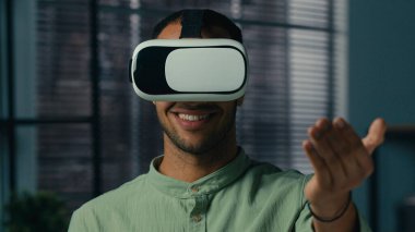 Close-up smiling latin american man wearing virtual reality glasses friendly showing invitation gesture asking to come welcome symbol making choice male player playing in cyberspace with vr goggles