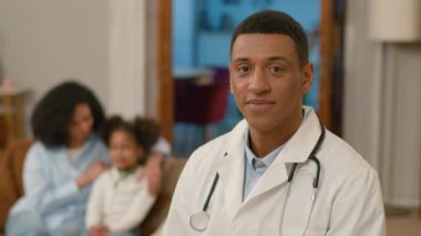 Afro-Amerikalı erkek doktor kameraya bakıyor gülümseyen konuşma terapisti çocuk doktoru ev sağlık görevlisi aile çalışanı anne ve kız hasta kızın tedavi odasına gitmesine yardım ediyor.